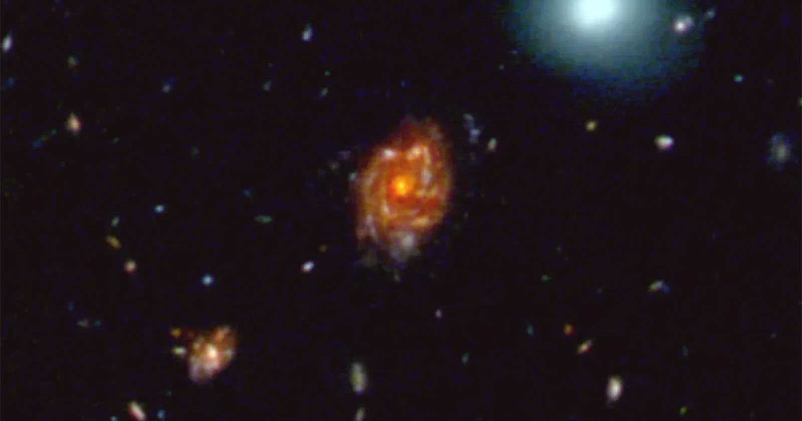 A deep-space image showing distant galaxies. The central spiral galaxy glows with bright orange and red hues, surrounded by smaller stars and galaxies against a black background.