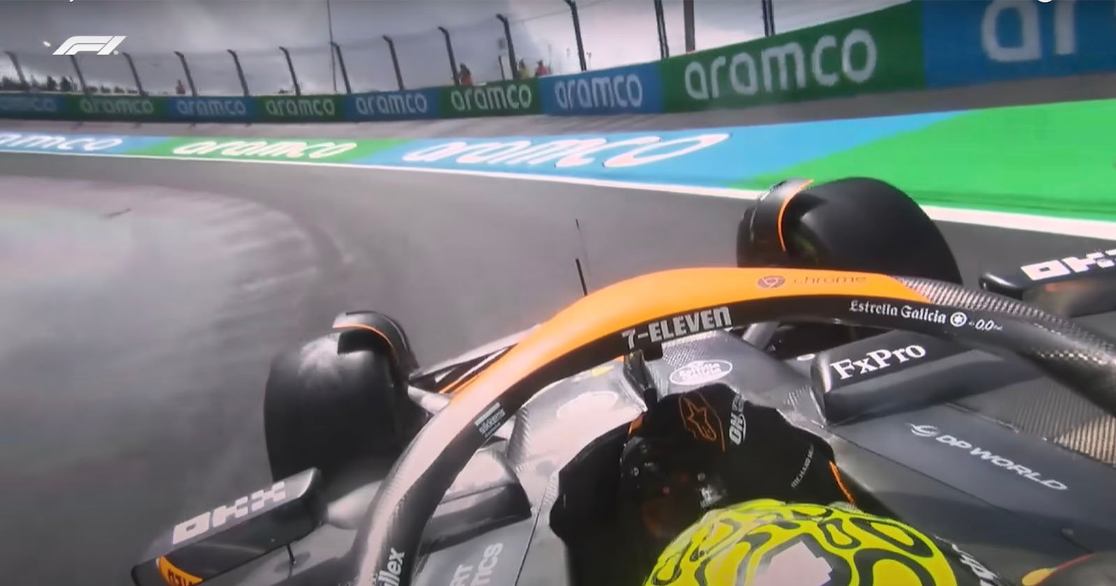 A close-up of a Formula 1 car from a first-person perspective, navigating a corner on a racetrack. The driver wears a brightly colored helmet, and sponsor logos, including 7-Eleven, are visible on the vehicle. The track is wet, and aramco branding is seen on barriers.