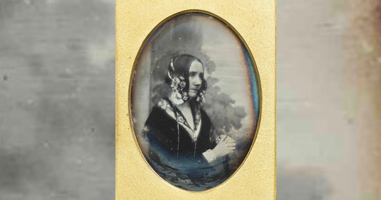 A sepia-toned portrait of a woman in Victorian-era clothing, with floral hair accessories, seated and looking to the side. The image is oval-shaped and framed in gold.