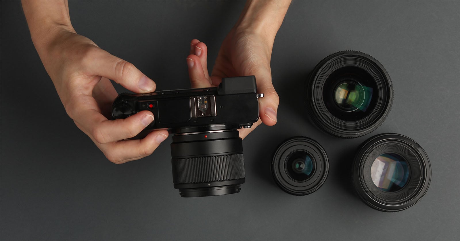A person holds a digital camera with a lens attached, while three additional camera lenses are arranged on a dark surface nearby.