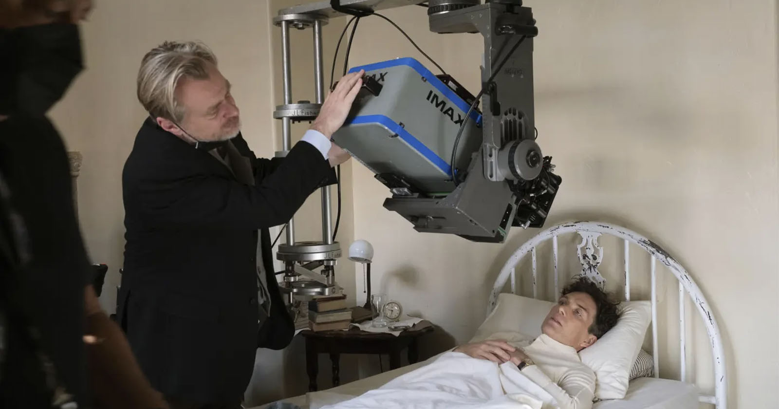 A person in a black suit and white shirt adjusts a large IMAX camera set up directly above another person lying in bed, who is covered with white sheets and has visible medical bandages. The scene is set in a room with a cream-colored wall and a vintage bed frame.