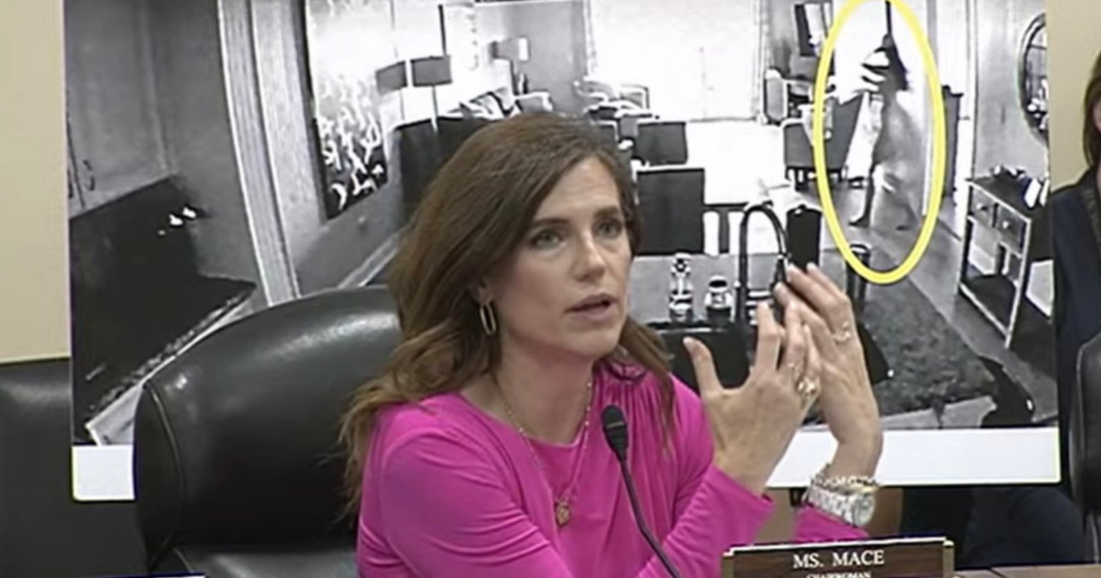 A woman in a pink top speaks at a meeting, gesturing with her hands. Behind her is a large black-and-white photo showing a living room and a figure highlighted in a yellow oval. A nameplate reads “Ms. Mace.”.