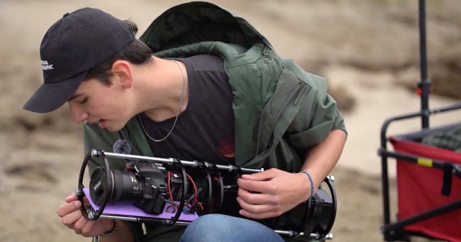 A person wearing a black cap and green jacket examines a cylindrical underwater camera housing while sitting outdoors on a sandy surface.