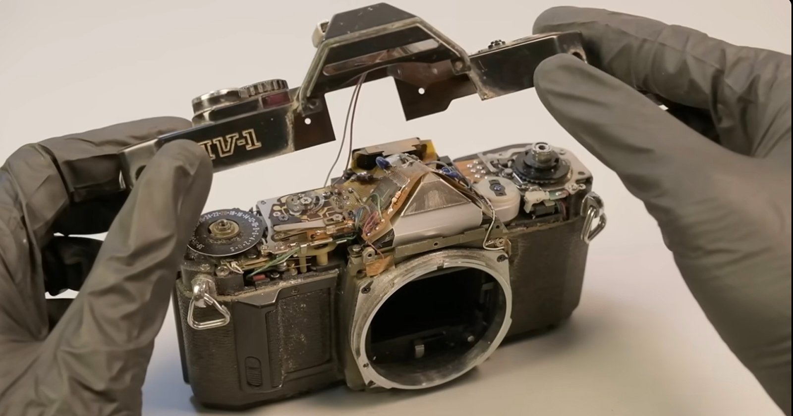 A person wearing grey gloves is disassembling a vintage film camera. The top section, labeled LV-1, is being carefully removed, exposing the mechanical components inside the camera body.