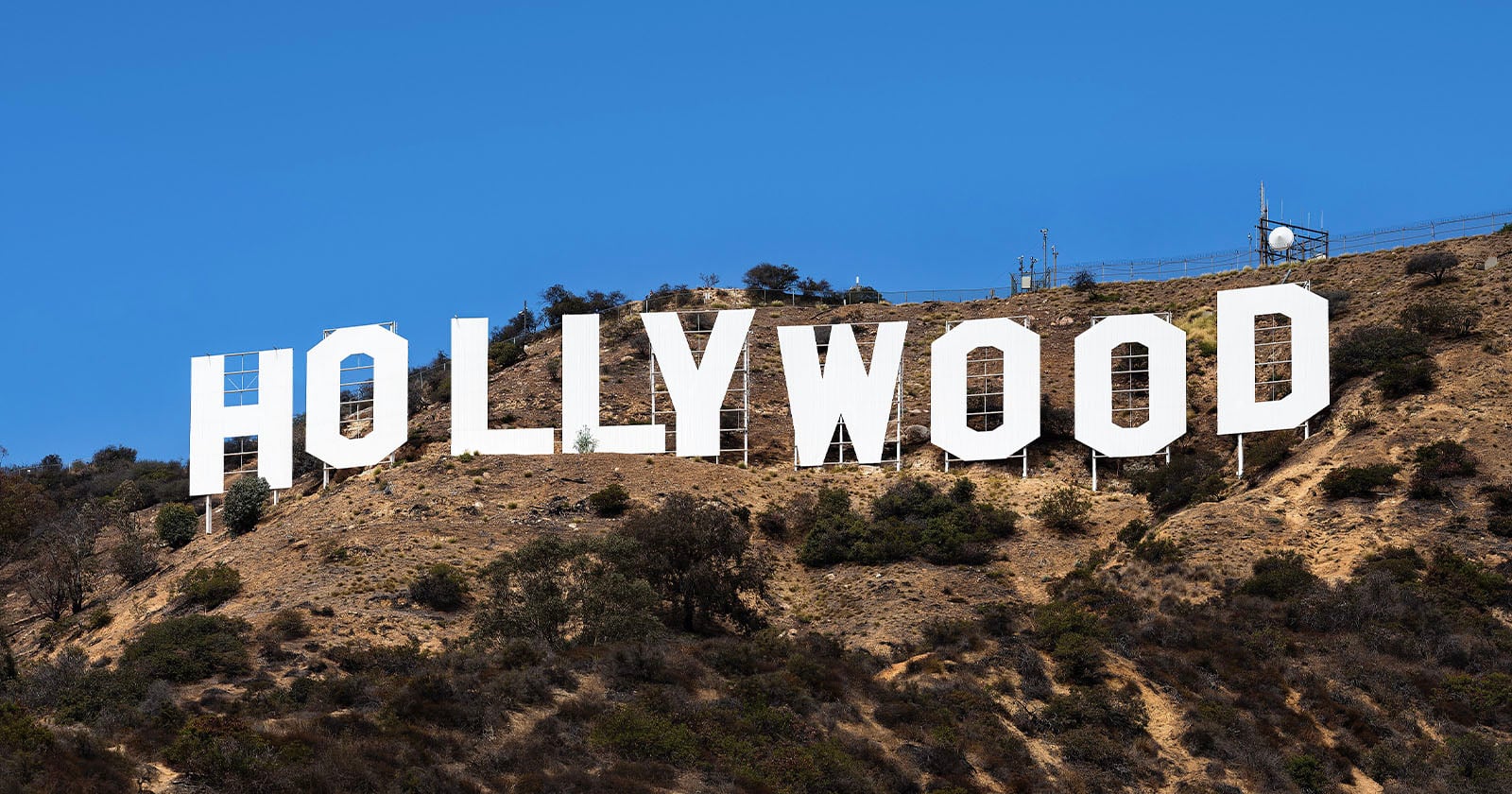 Large white letters spelling HOLLYWOOD stand on a dry, shrub-covered hillside under a clear blue sky. The famous landmark is set against rugged terrain with sparse vegetation.
