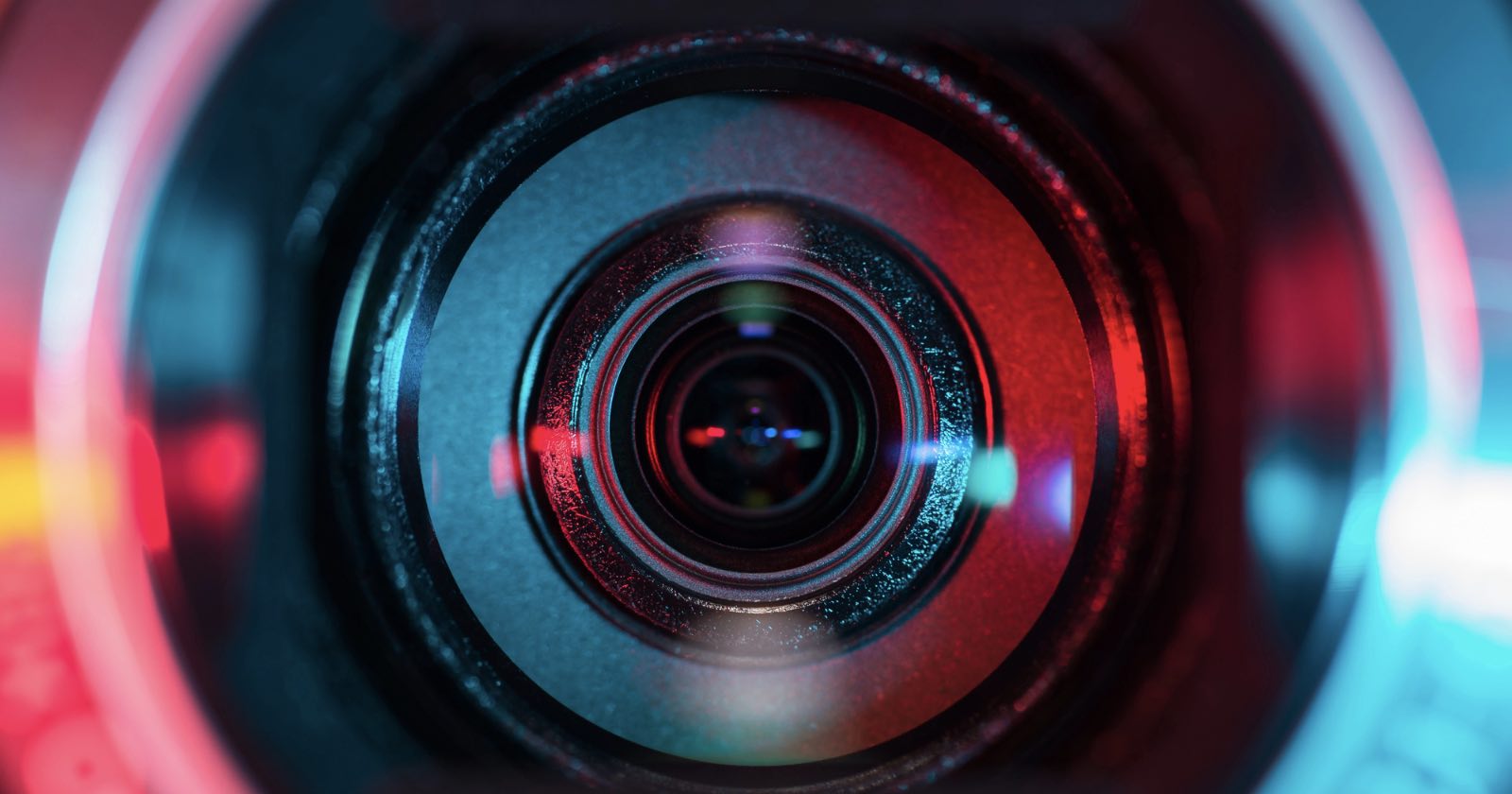 Close-up image of a camera lens, with reflections and colorful lighting in shades of red and blue highlighting the lens rings and glass.