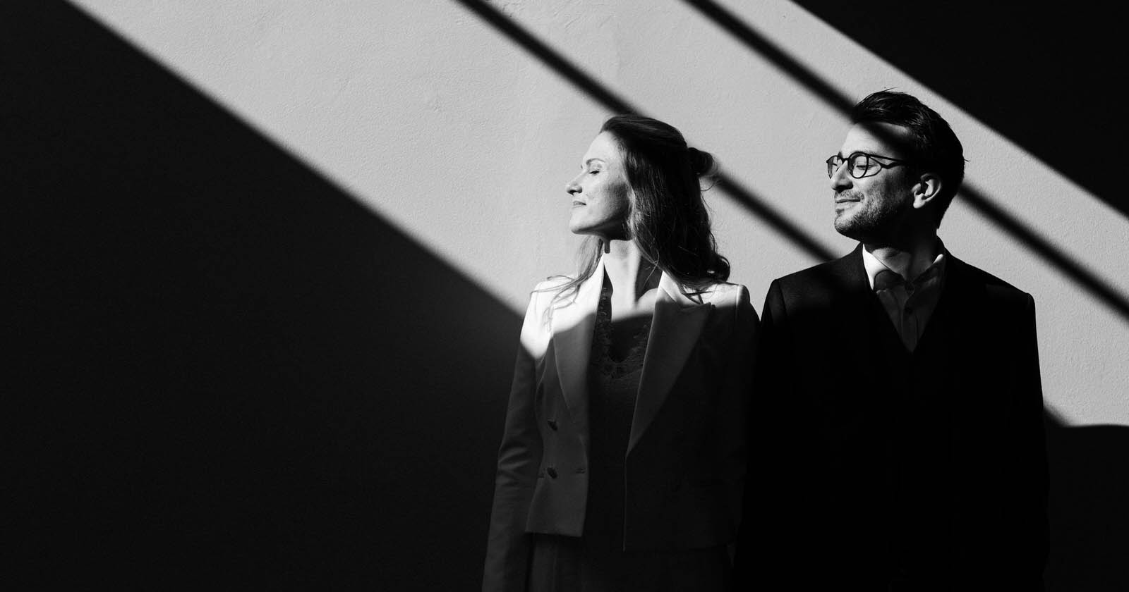 A black and white image of a man and a woman in business attire standing side by side against a wall, with defined shadows from overhead light casting lines over them.