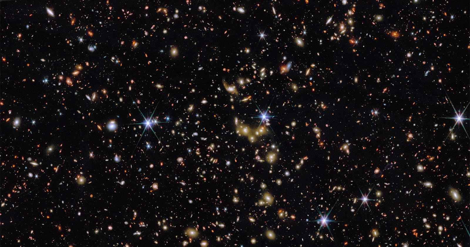 A deep space image showing countless distant galaxies, stars, and bright points of light scattered across a black background, with some stars displaying diffraction spikes.