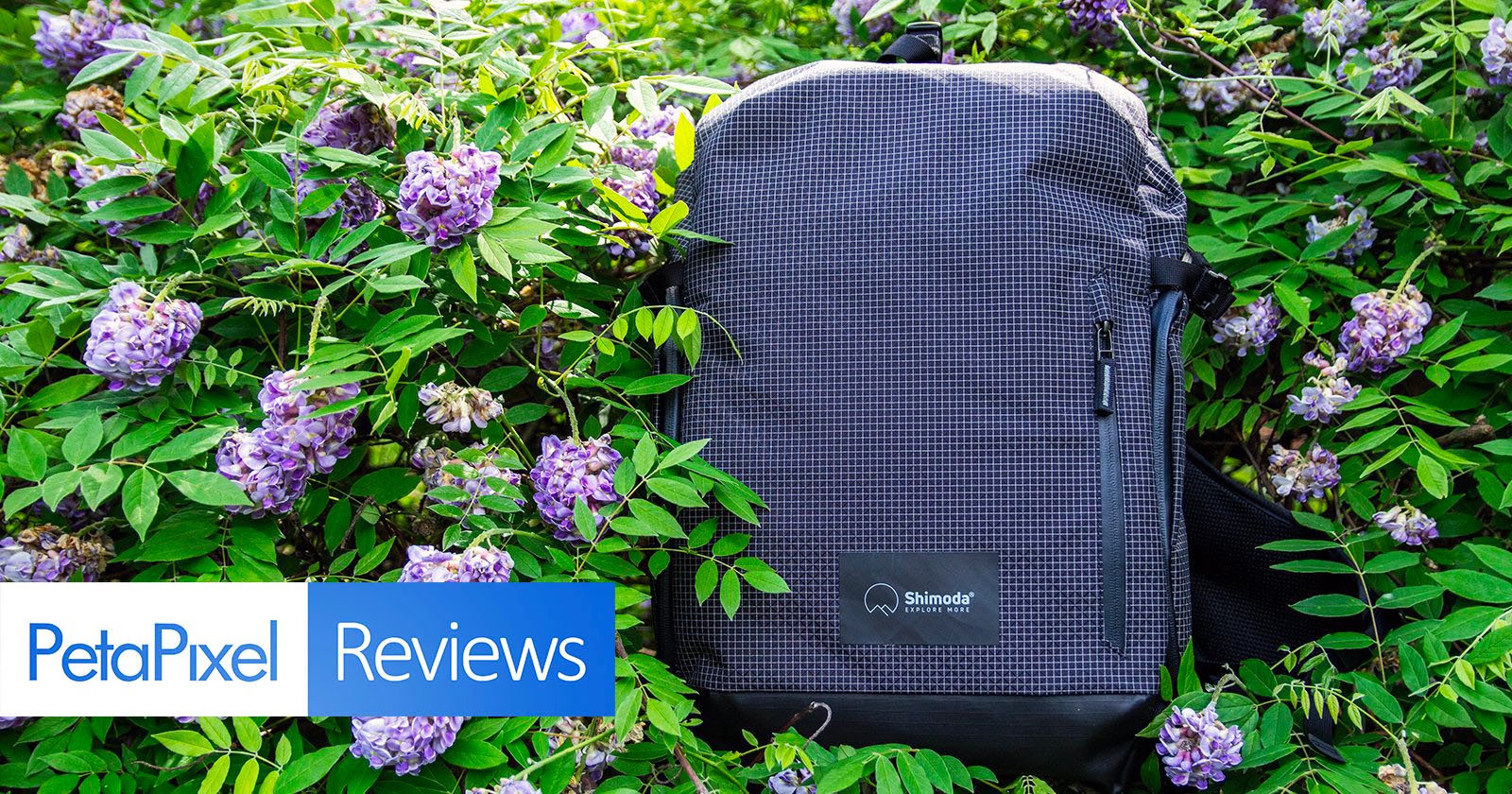 A black, grid-patterned Shimoda backpack sits among lush green leaves and purple flowers. Overlay text reads PetaPixel Reviews in the lower left corner.