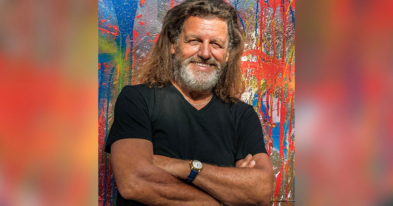 A man with long hair and a beard smiles, standing with arms crossed. He wears a black t-shirt and a watch, against a colorful abstract painted background.