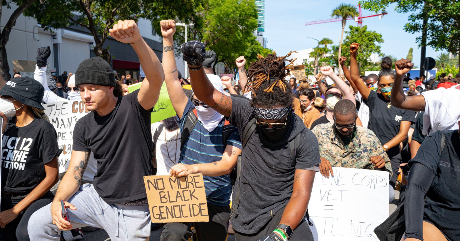 A group of protesters, diverse in age and ethnicity, kneels in solidarity with their fists raised. Some hold signs with messages such as No More Black Genocide and Marine Corps Vet = Willing to Die. The atmosphere is tense and passionate.