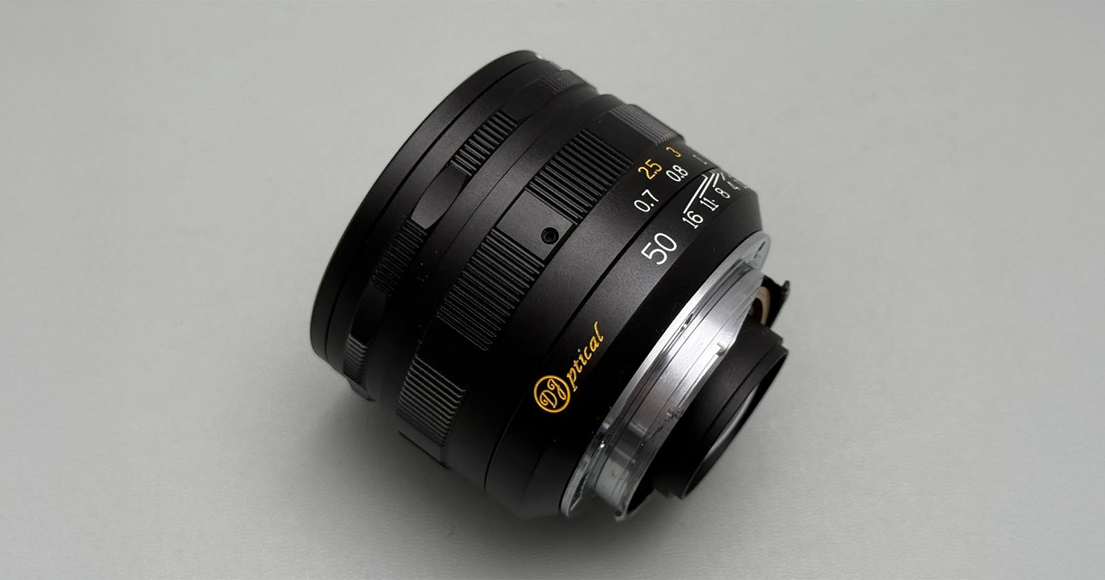 A close-up photo of a black camera lens with gold and white markings, including 50 for the focal length, set against a plain light gray background.