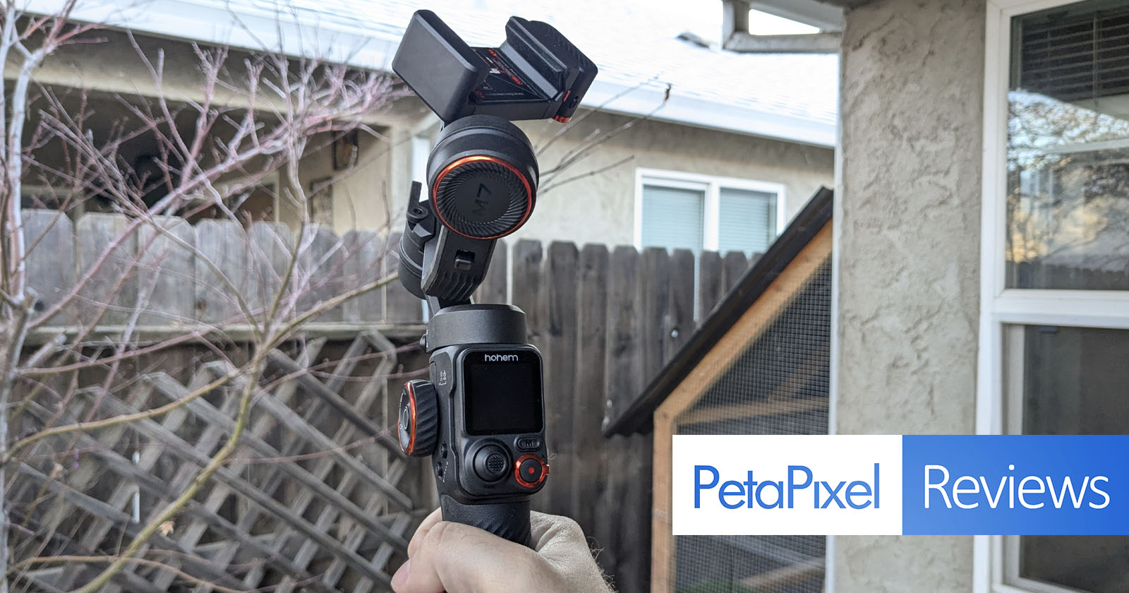 A person holds a Hohem camera gimbal outdoors near a wooden fence. The gimbal features a screen and buttons and is positioned against a backdrop of a house and leafless branches. The image includes a PetaPixel Reviews logo.