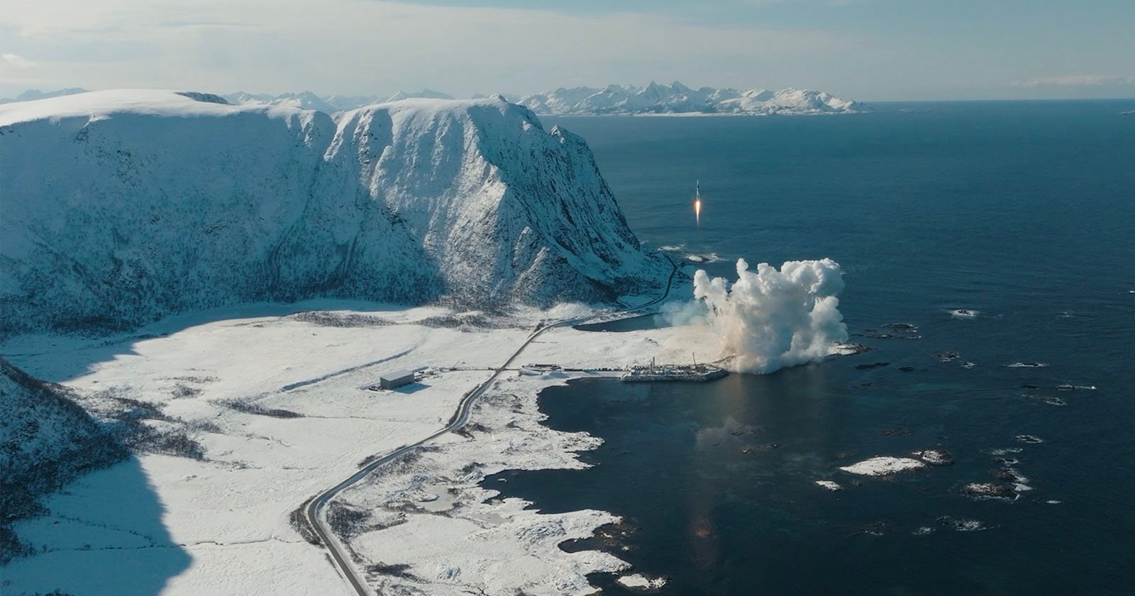 A rocket launches from a snowy coastal area surrounded by mountains and ocean. Smoke and flames are visible from the launch site. The landscape is covered in snow, and the sea is calm under a clear sky.