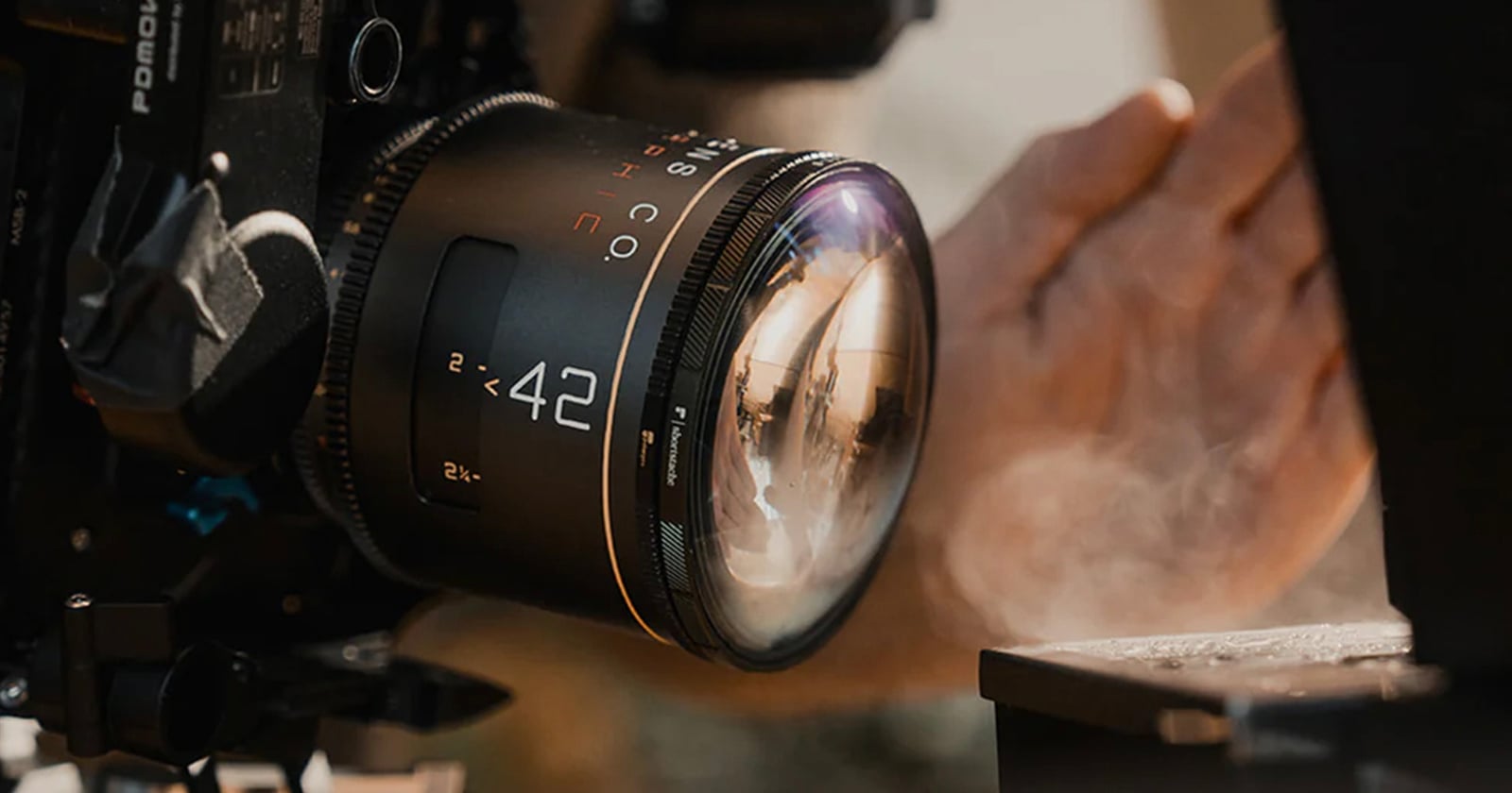 Close-up of a camera with a large 42mm lens in focus, capturing a scene. A hand is visible in the background, partially blurred, with sunlight reflecting off the lens and a bit of smoke or mist rising nearby.