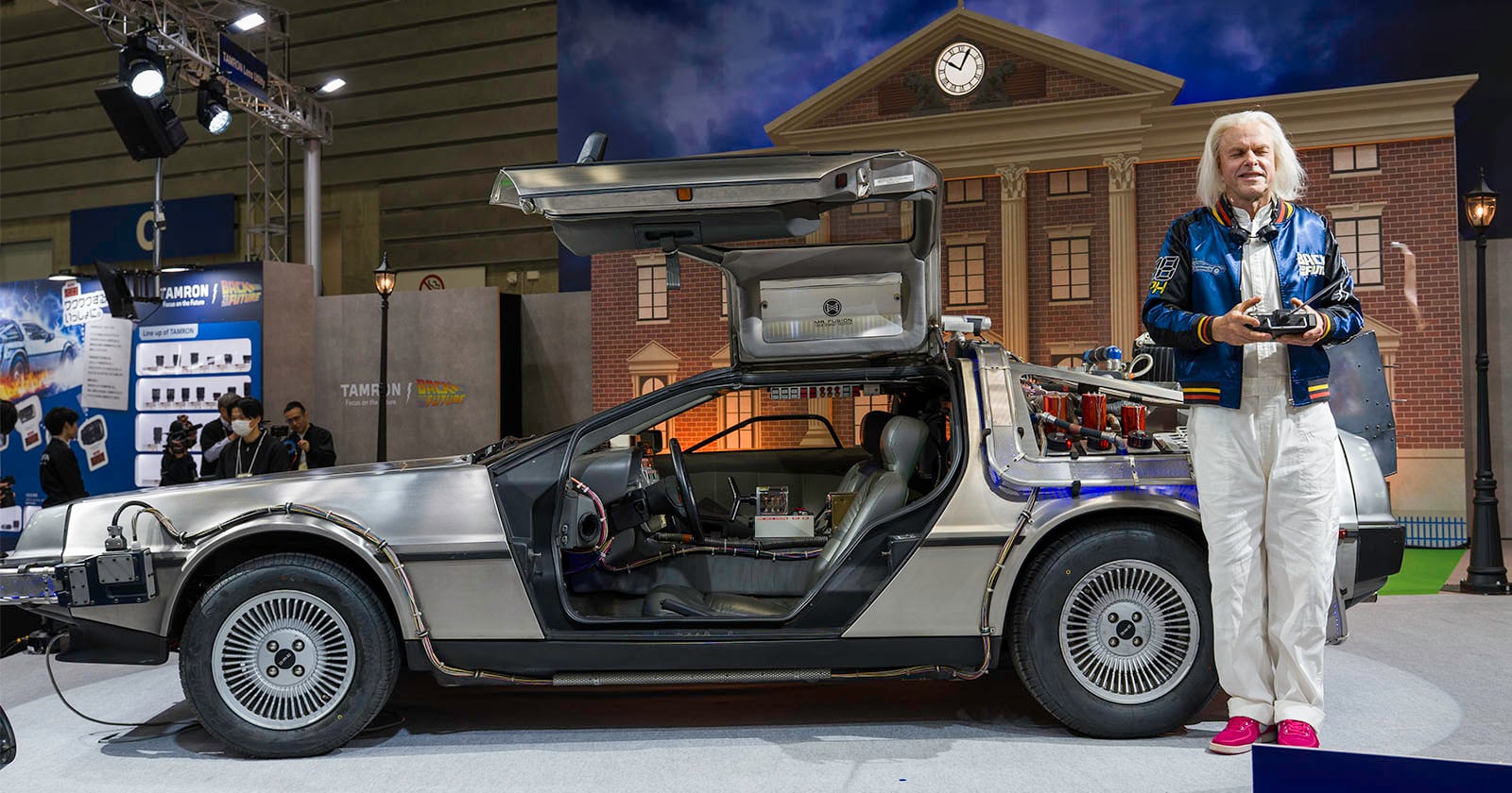 A man with white hair, wearing a blue jacket and white pants, stands next to a DeLorean car with gull-wing doors open, in front of a backdrop resembling the Back to the Future clock tower.