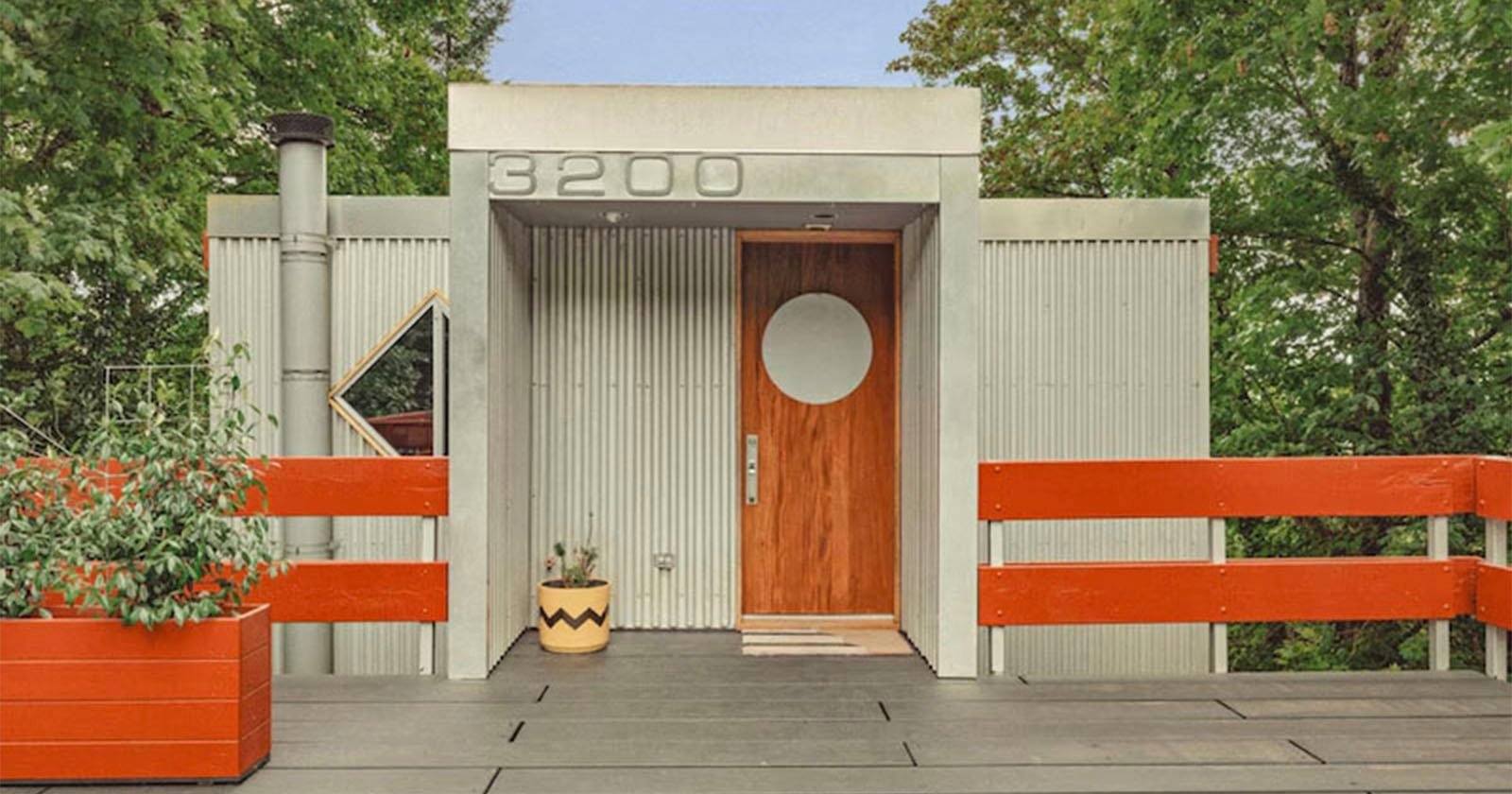 Modern house entrance with gray corrugated metal exterior, a wooden door featuring a circular window, red railings, large house number 3200 above, and potted plants by the door, surrounded by green trees.