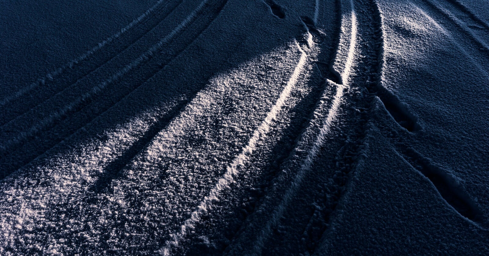 Tire tracks and footprints are pressed into a snowy surface, illuminated by soft, angled sunlight that casts contrasting shadows and highlights on the textured snow.