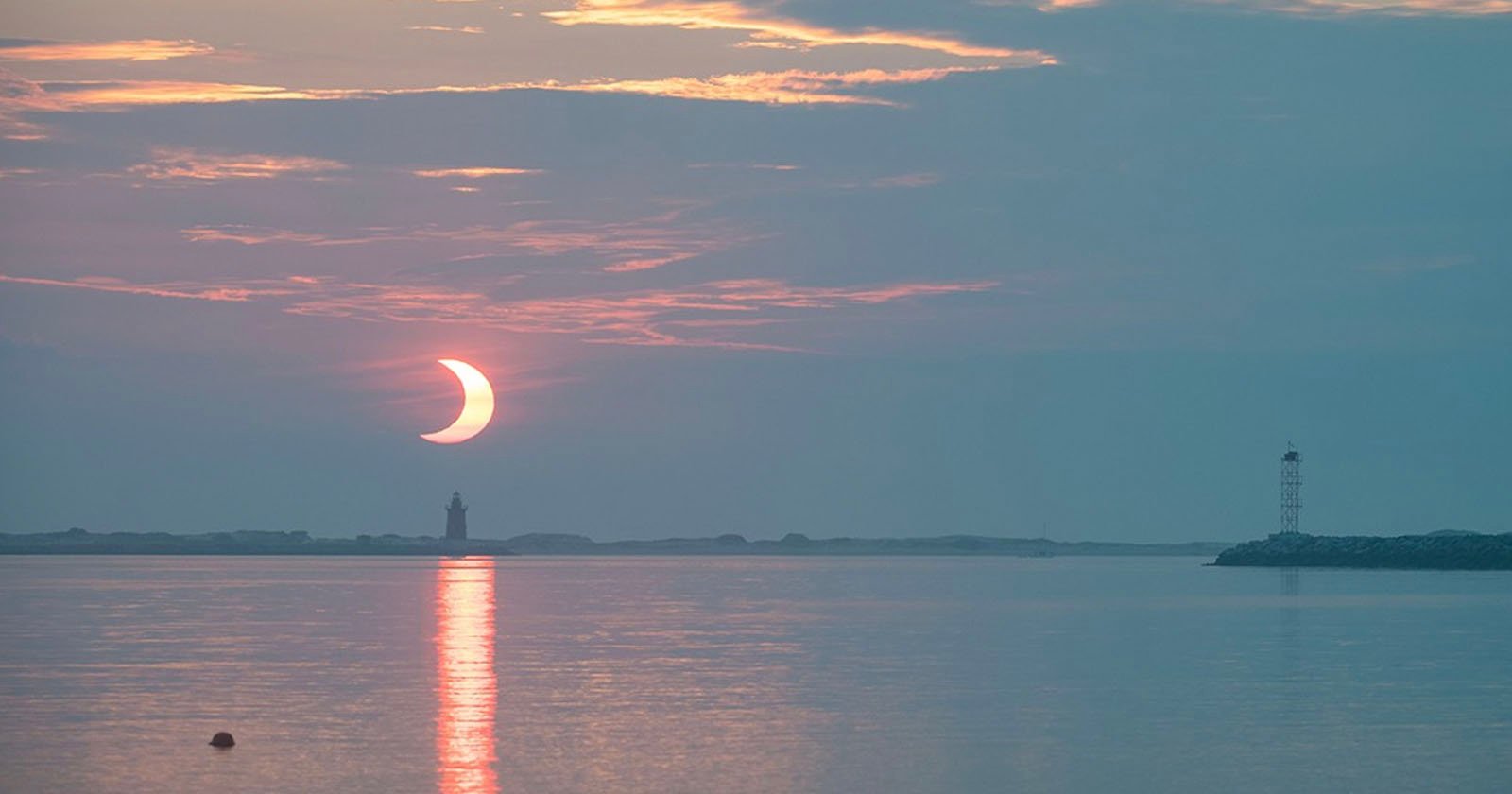 The image depicts a partial solar eclipse over calm water at sunrise or sunset. The sky is a mix of soft pink and blue hues. A lighthouse and small tower are visible in silhouette against the horizon.