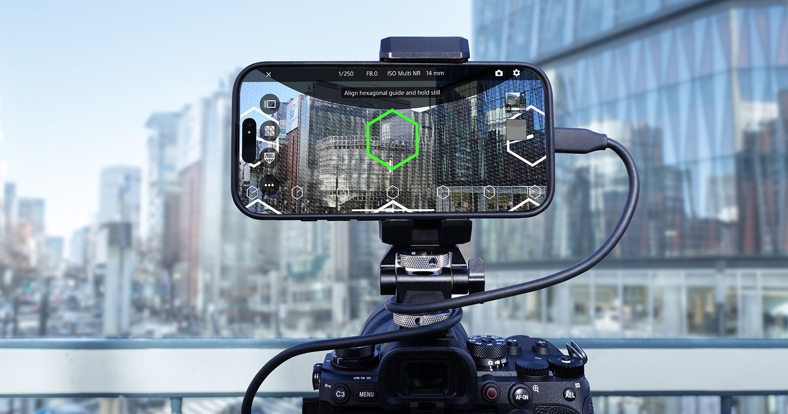 A smartphone mounted on a camera is displaying a cityscape with a digital overlay of hexagonal guides. Both devices are set up on a balcony with tall glass buildings in the background.