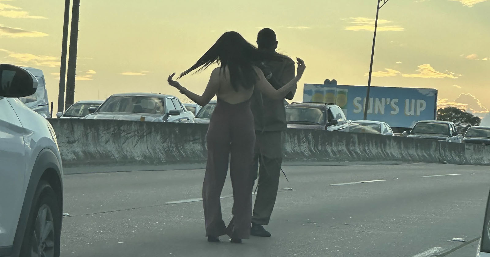 Two people are walking on a highway between lanes of slow-moving traffic. The person on the left is flipping their long hair, and the person on the right is wearing a hoodie. A billboard in the background says SUN'S UP.