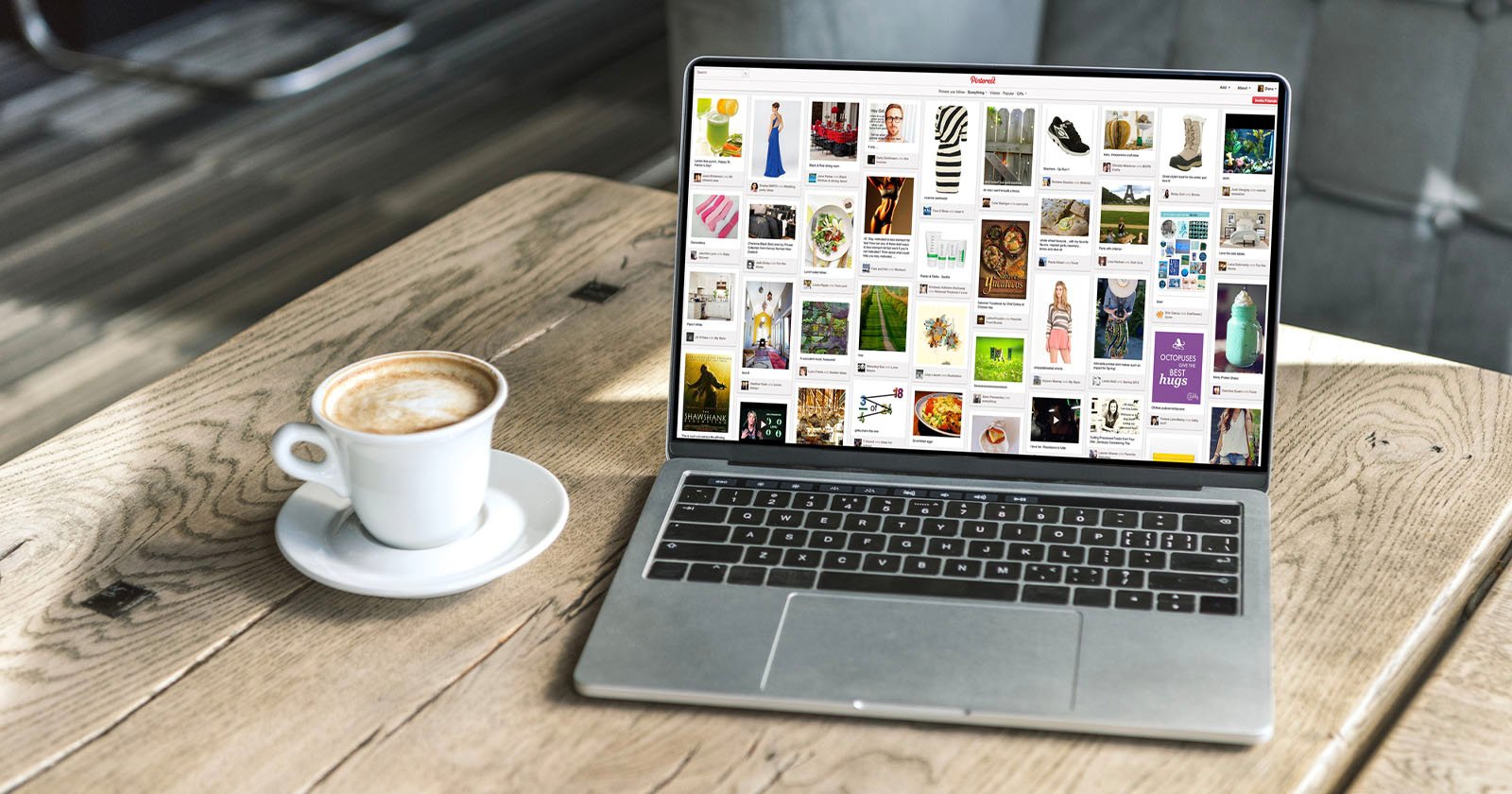A laptop displaying a Pinterest page with various image pins on its screen is placed on a wooden table. Beside it, there is a cup of cappuccino. The background shows a blurred indoor setting with a grey chair.