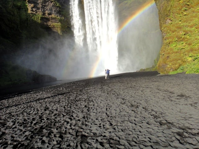 skogafoss-waterfall-black-pebbly-2-tiny-people-in-front-of-bright-water-falling-arc-of-rainbow-between-water-and-rockface-upper-right.jpg