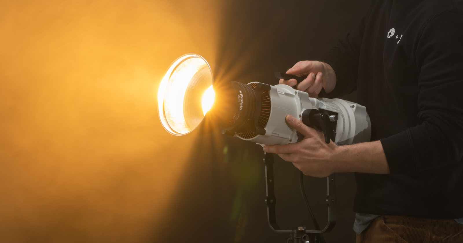 A person holding an LED light with a large attached reflector, aiming a bright light towards the left, in a studio with a warm, orange-hued backdrop.