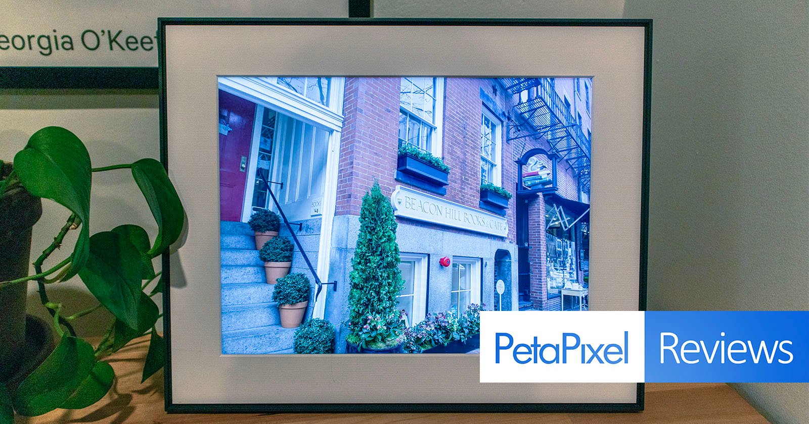 A digital frame displaying a photo of a quaint blue building with a sign labeled pelton place. the frame is on a wooden table, flanked by a potted plant and a book titled georgia o'keefe.