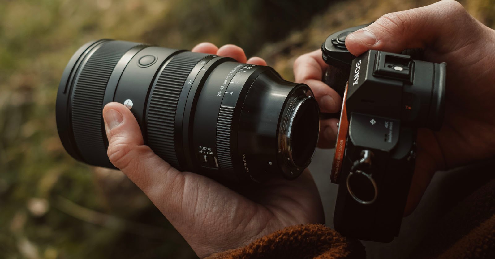 A person is holding a Sony camera in their right hand and a detached Sigma lens in their left hand. The background appears to be an outdoor setting with greenery. A person is holding a Sony camera in their right hand and a detached Sigma lens in their left hand. The background appears to be an outdoor setting with greenery.