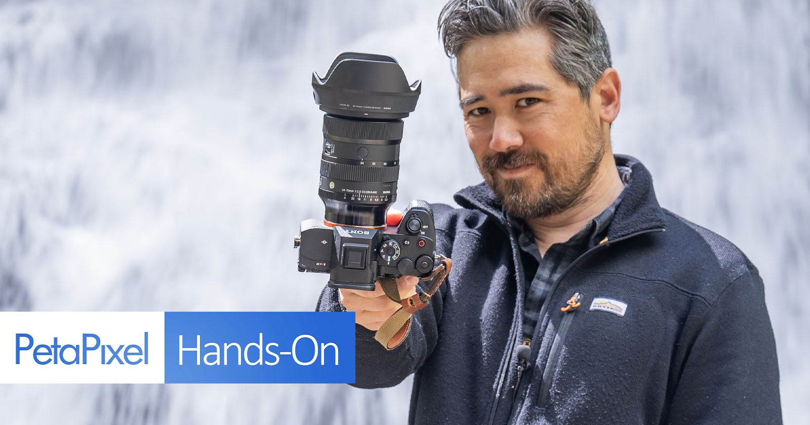 A man holding a professional camera with a large lens is standing in front of a waterfall. He is wearing a dark jacket, and there is a PetaPixel Hands-On graphic at the bottom left corner of the image. A man holding a professional camera with a large lens is standing in front of a waterfall. He is wearing a dark jacket, and there is a PetaPixel Hands-On graphic at the bottom left corner of the image.