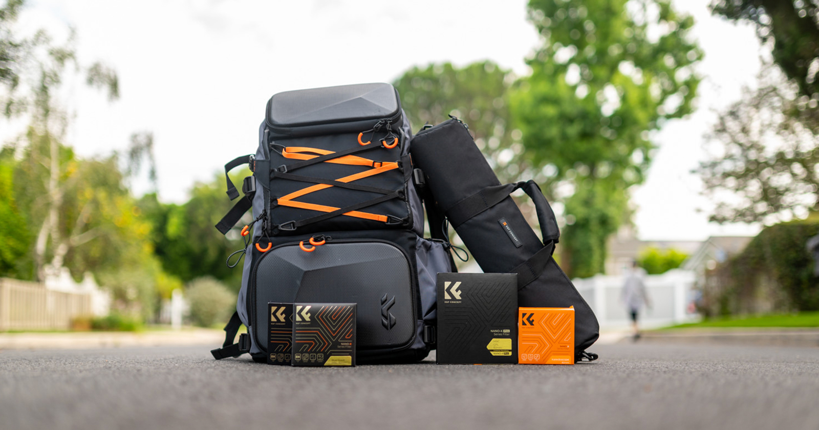 An assortment of outdoor gear is displayed on the road, including two black and orange backpacks, a black cylindrical bag, and three orange and black boxes with a logo and text. Background shows trees and houses, with a person walking in the distance. An assortment of outdoor gear is displayed on the road, including two black and orange backpacks, a black cylindrical bag, and three orange and black boxes with a logo and text. Background shows trees and houses, with a person walking in the distance.