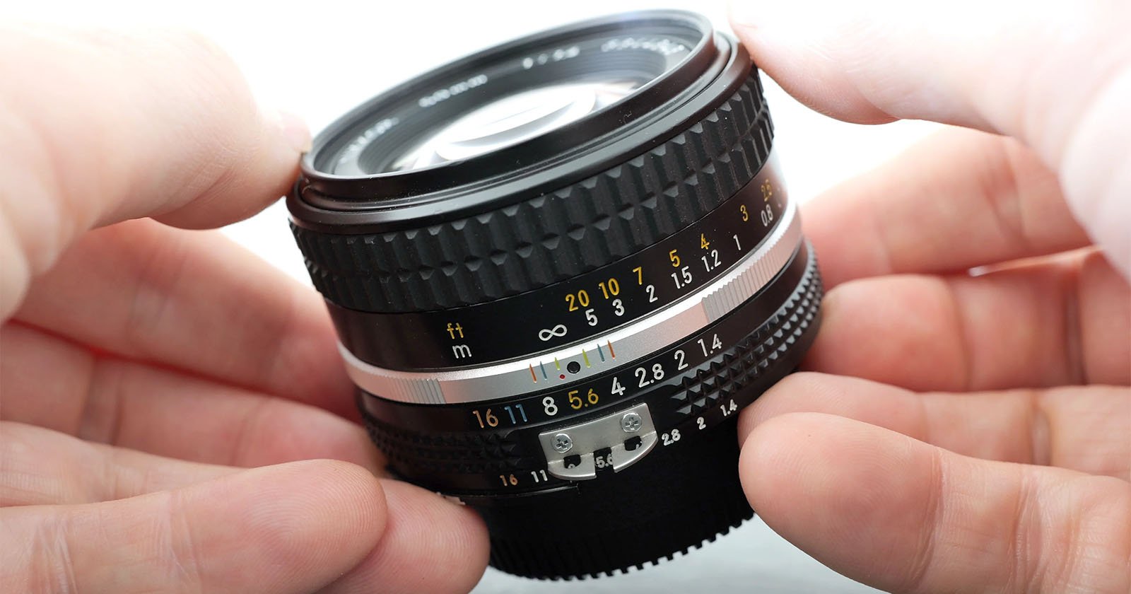 Close-up of hands holding a camera lens. The lens features various markings, including numbers representing aperture settings (from f/1.4 to f/16) and distance measurements in meters and feet. The focus ring and other detailed components are prominently visible. Close-up of hands holding a camera lens. The lens features various markings, including numbers representing aperture settings (from f/1.4 to f/16) and distance measurements in meters and feet. The focus ring and other detailed components are prominently visible.