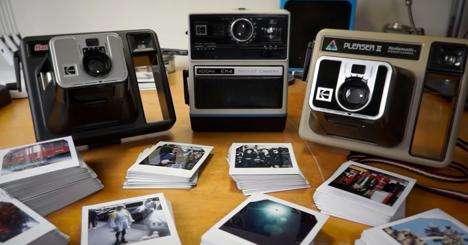 A collection of three vintage Kodak instant cameras are displayed on a wooden table surrounded by piles of Polaroid photos. The camera models include the EK4, The Handle, and the Pleaser II. The photos depict various scenes and are arranged neatly in front. A collection of three vintage Kodak instant cameras are displayed on a wooden table surrounded by piles of Polaroid photos. The camera models include the EK4, The Handle, and the Pleaser II. The photos depict various scenes and are arranged neatly in front.
