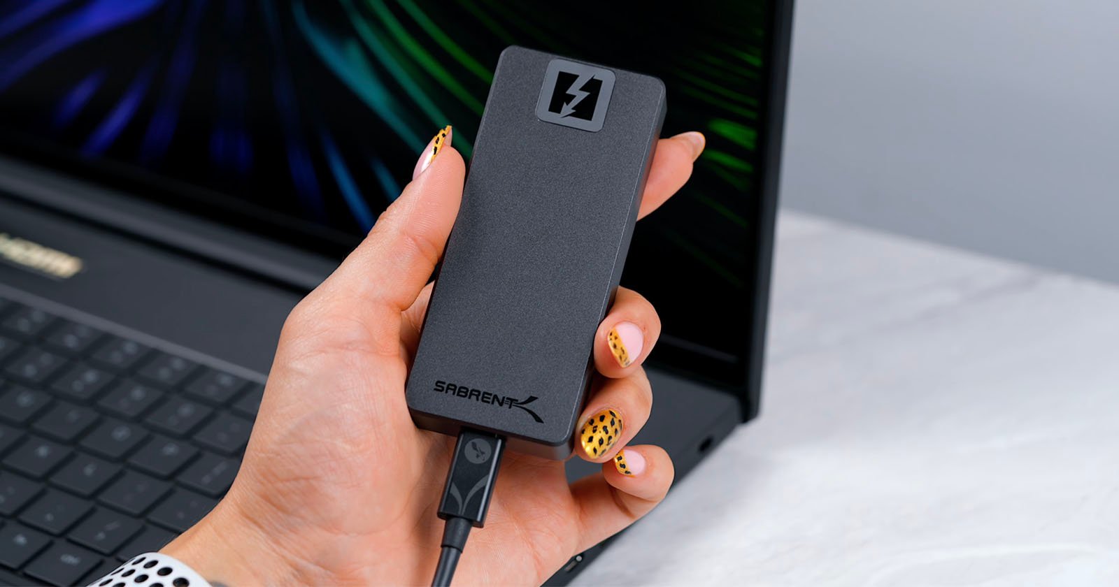 A hand with decorated nails holds a black Sabrent external SSD, which has a lightning bolt icon. The SSD is connected to a laptop in the background. A hand with decorated nails holds a black Sabrent external SSD, which has a lightning bolt icon. The SSD is connected to a laptop in the background.