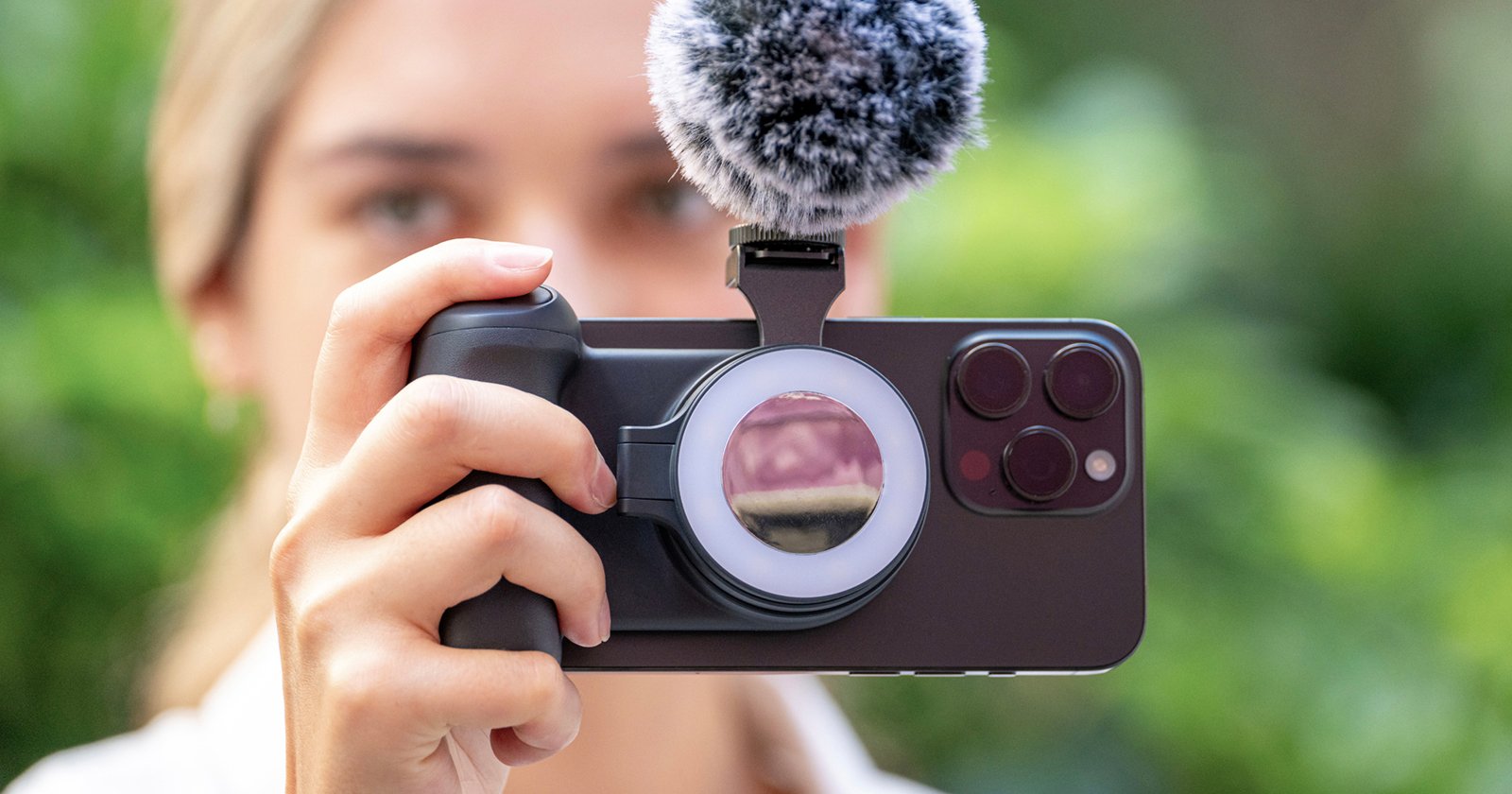 A person holds a smartphone with a microphone and a ring light attachment, pointing it toward the viewer. The phone has three visible camera lenses, and the hand gripping the phone has light-colored skin. The background is blurred greenery. A person holds a smartphone with a microphone and a ring light attachment, pointing it toward the viewer. The phone has three visible camera lenses, and the hand gripping the phone has light-colored skin. The background is blurred greenery.