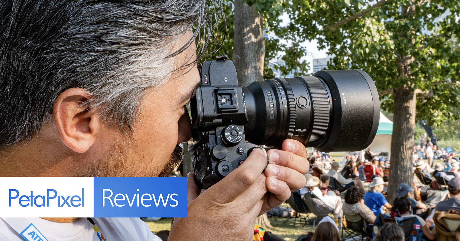 A person with short gray hair is closely holding and looking through a professional camera. The scene is outdoors with trees and a crowd in the background. The image has banners with the text PetaPixel and Reviews. A person with short gray hair is closely holding and looking through a professional camera. The scene is outdoors with trees and a crowd in the background. The image has banners with the text PetaPixel and Reviews.