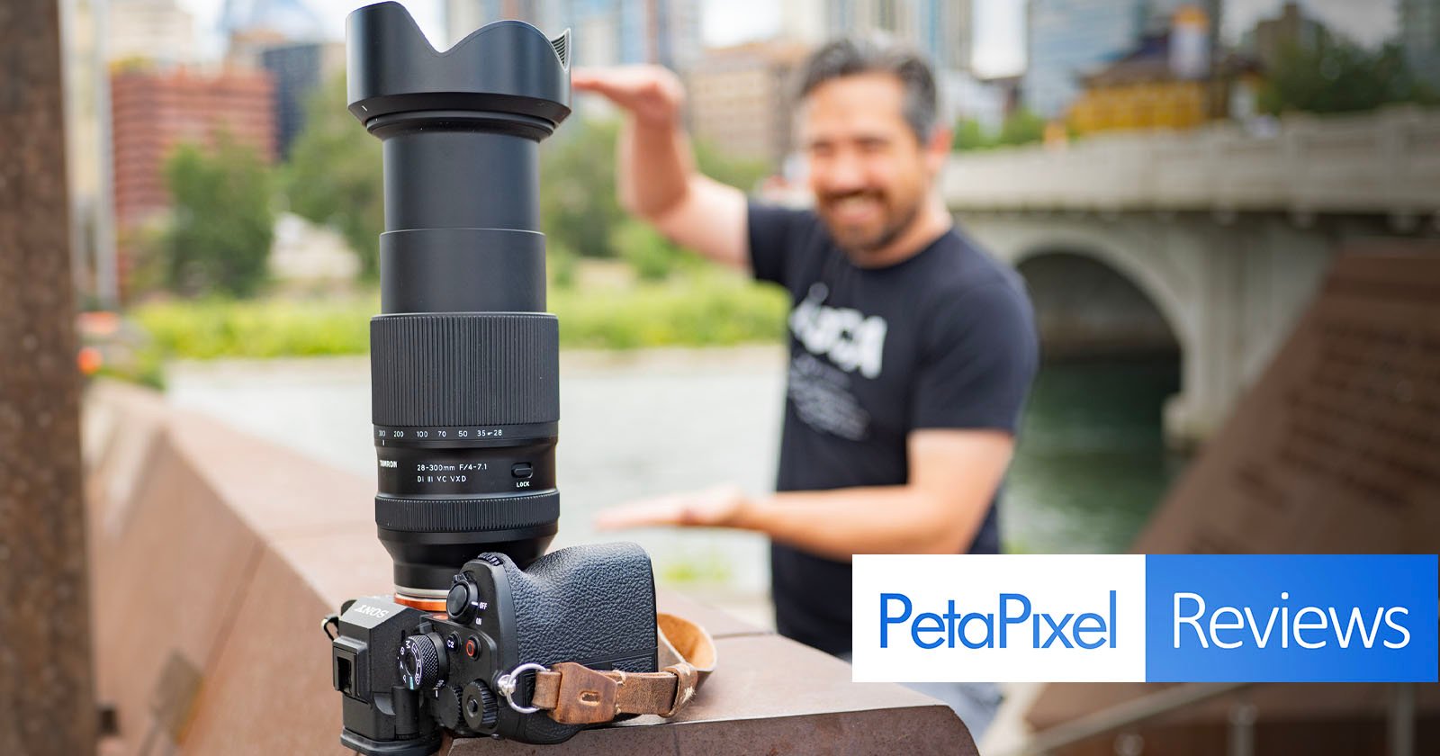 A camera with a large lens is placed on a ledge outdoors, with an arm strap attached. In the blurred background, a smiling man stands next to a river and bridge, holding his hands out as if framing the shot. The image is overlaid with the logo and text PetaPixel Reviews. A camera with a large lens is placed on a ledge outdoors, with an arm strap attached. In the blurred background, a smiling man stands next to a river and bridge, holding his hands out as if framing the shot. The image is overlaid with the logo and text PetaPixel Reviews.