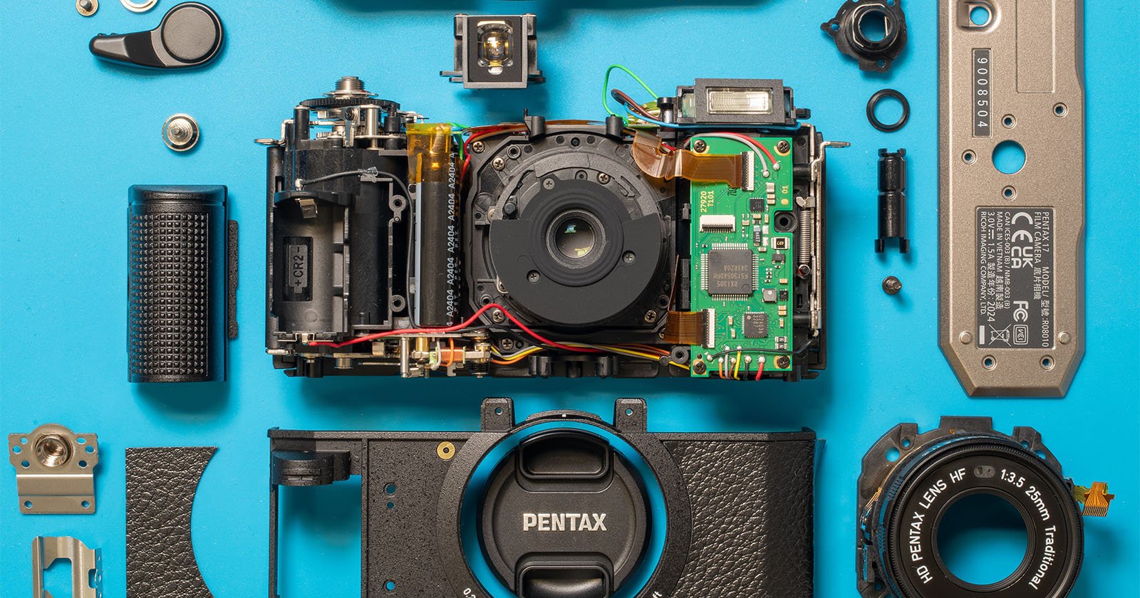 Top-down view of a disassembled camera with various internal components, including the lens, circuitry, screws, and body parts, laid out on a blue background. The brand name Pentax is visible on some of the parts. Top-down view of a disassembled camera with various internal components, including the lens, circuitry, screws, and body parts, laid out on a blue background. The brand name Pentax is visible on some of the parts.