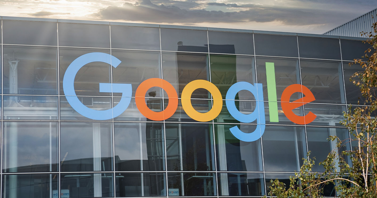 A large sign displaying the word Google in colorful letters is mounted on the glass facade of a modern building. The background includes a sky with clouds and some sunlight. A tree with green leaves is visible in the bottom right corner. A large sign displaying the word Google in colorful letters is mounted on the glass facade of a modern building. The background includes a sky with clouds and some sunlight. A tree with green leaves is visible in the bottom right corner.