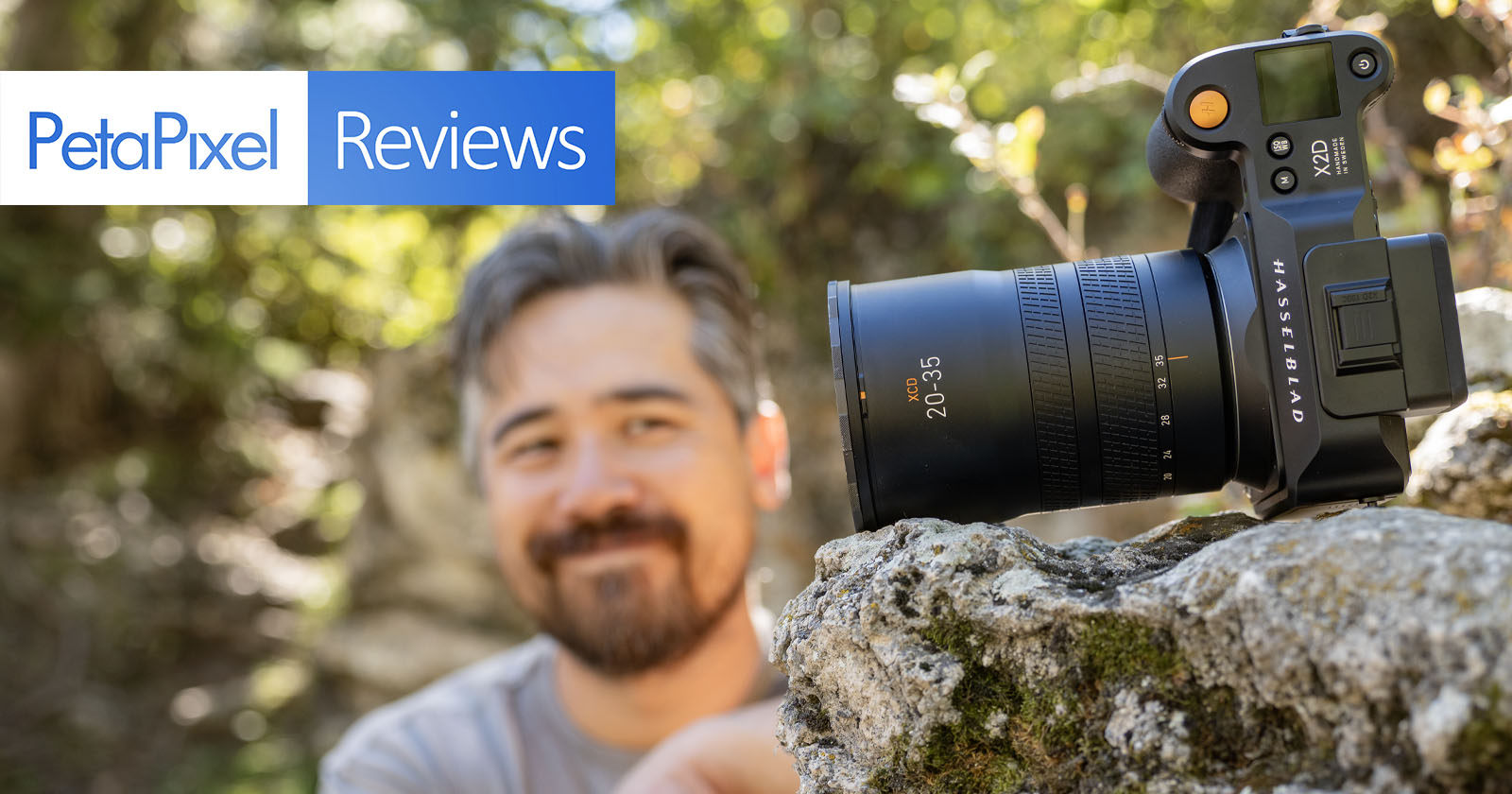 A camera with a large lens is perched on a rock outdoors. In the background, a person with a beard and mustache is slightly out of focus, smiling and looking at the camera. The text PetaPixel Reviews is displayed in the upper left corner. A camera with a large lens is perched on a rock outdoors. In the background, a person with a beard and mustache is slightly out of focus, smiling and looking at the camera. The text PetaPixel Reviews is displayed in the upper left corner.