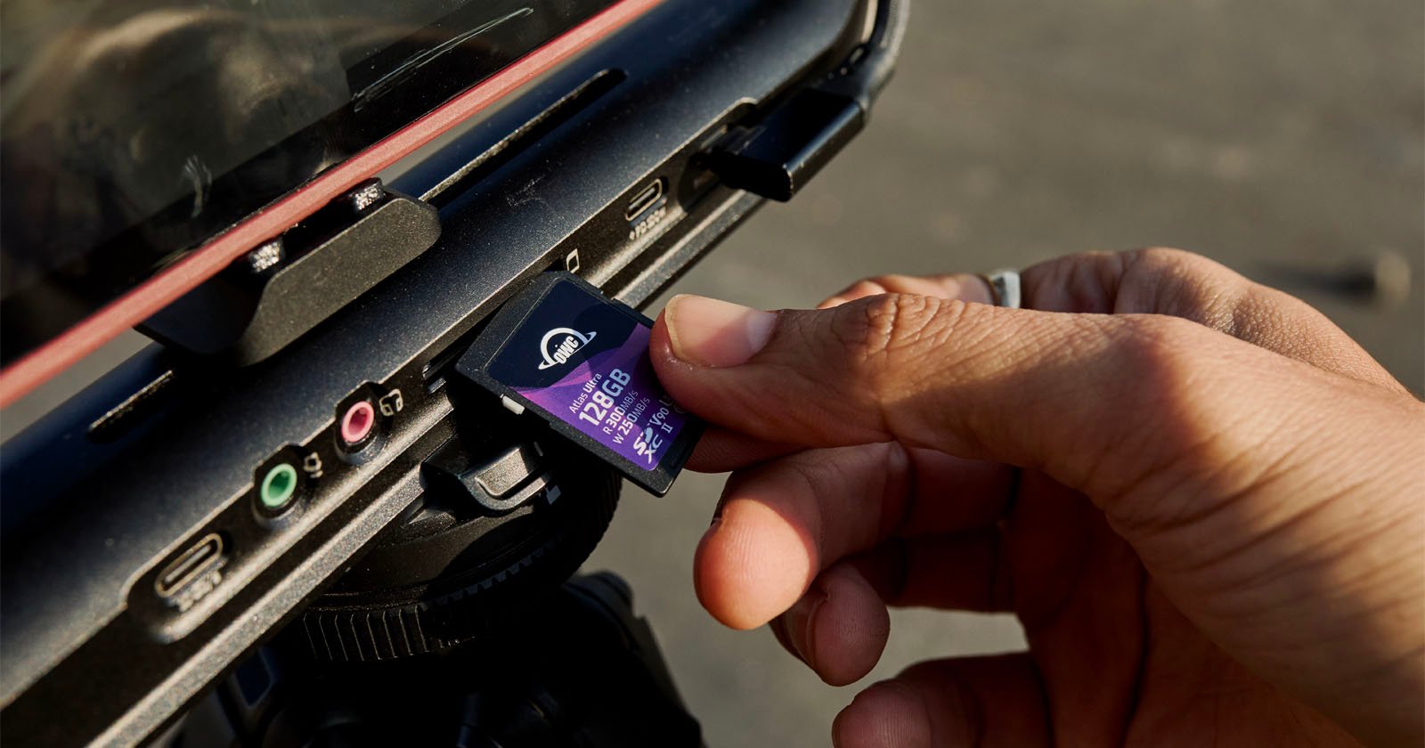 A close-up image of a person's hand inserting a purple 128GB SD card into a slot on the side of a camera or similar device. The person's fingers are holding the edge of the card, and different input ports are visible around the slot area. A close-up image of a person's hand inserting a purple 128GB SD card into a slot on the side of a camera or similar device. The person's fingers are holding the edge of the card, and different input ports are visible around the slot area.