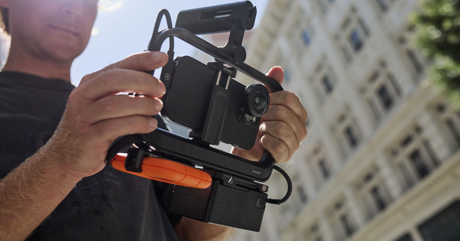 A person holds a professional video rig with a mounted smartphone and microphone setup, possibly for filming. In the background, there is a tall building and some greenery, indicating an outdoor urban setting. A person holds a professional video rig with a mounted smartphone and microphone setup, possibly for filming. In the background, there is a tall building and some greenery, indicating an outdoor urban setting.