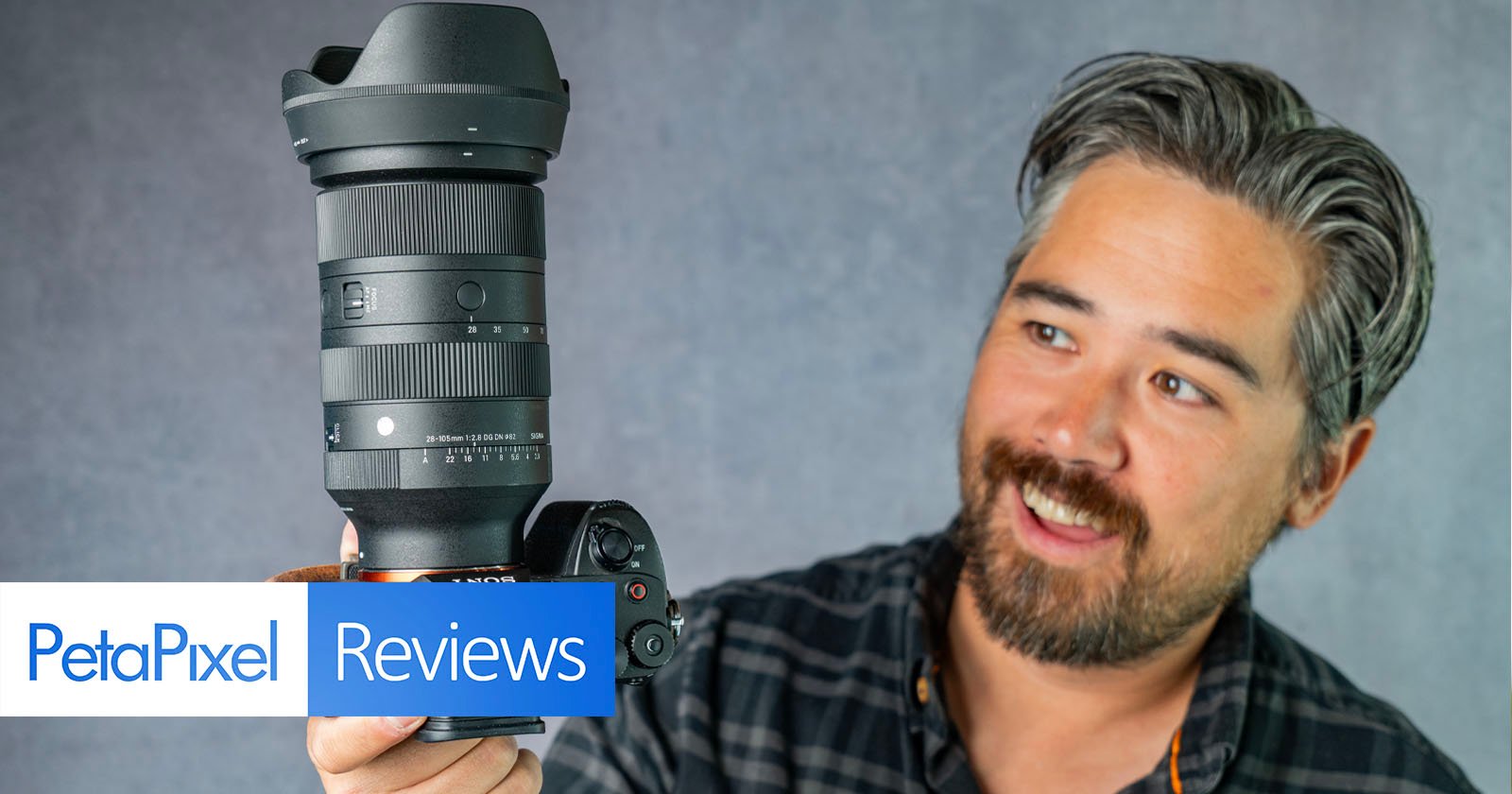 A man with gray-streaked hair and a beard smiles while holding up a DSLR camera with a large lens against a gray background. A banner at the bottom reads PetaPixel Reviews. A man with gray-streaked hair and a beard smiles while holding up a DSLR camera with a large lens against a gray background. A banner at the bottom reads PetaPixel Reviews.