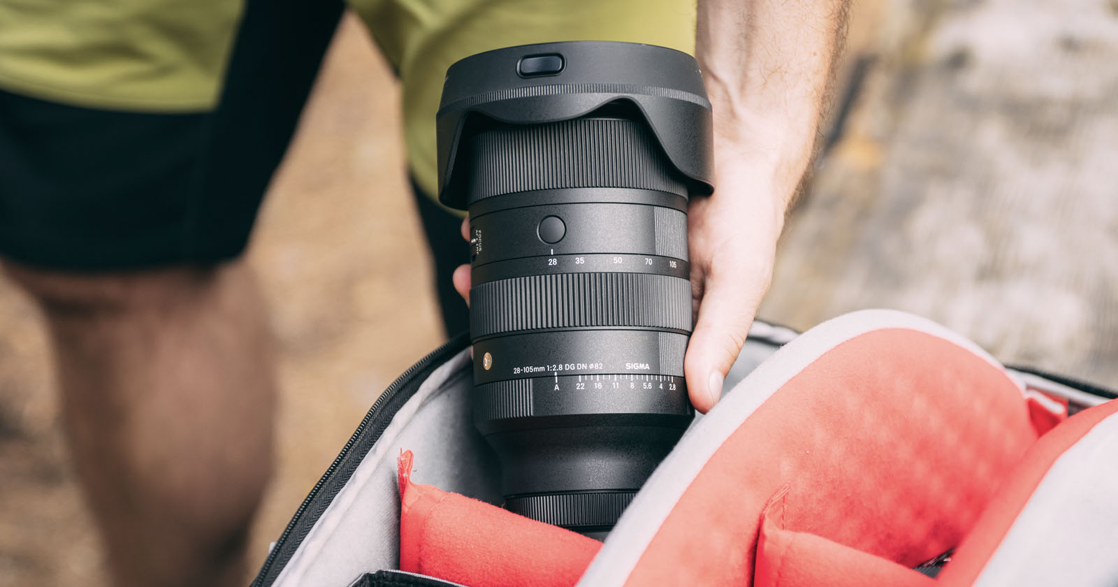 A person is holding a Sigma camera lens above an open camera bag with a red interior. The lens is black with various markings and a lens hood attached. The scene takes place outdoors, as indicated by the background. A person is holding a Sigma camera lens above an open camera bag with a red interior. The lens is black with various markings and a lens hood attached. The scene takes place outdoors, as indicated by the background.