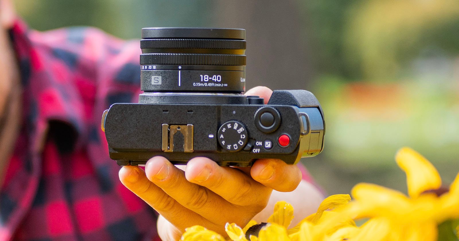 Person holding a black camera with an 18-40mm lens, surrounded by yellow flowers in the foreground. The person is wearing a red plaid shirt, and a blurred outdoor background is visible. Person holding a black camera with an 18-40mm lens, surrounded by yellow flowers in the foreground. The person is wearing a red plaid shirt, and a blurred outdoor background is visible.