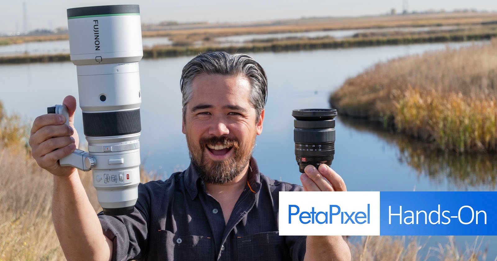 A smiling man outdoors holds a large camera lens in one hand and a smaller lens in the other. He stands near a water body with grass and reeds. The image includes a PetaPixel Hands-On logo. A smiling man outdoors holds a large camera lens in one hand and a smaller lens in the other. He stands near a water body with grass and reeds. The image includes a PetaPixel Hands-On logo.