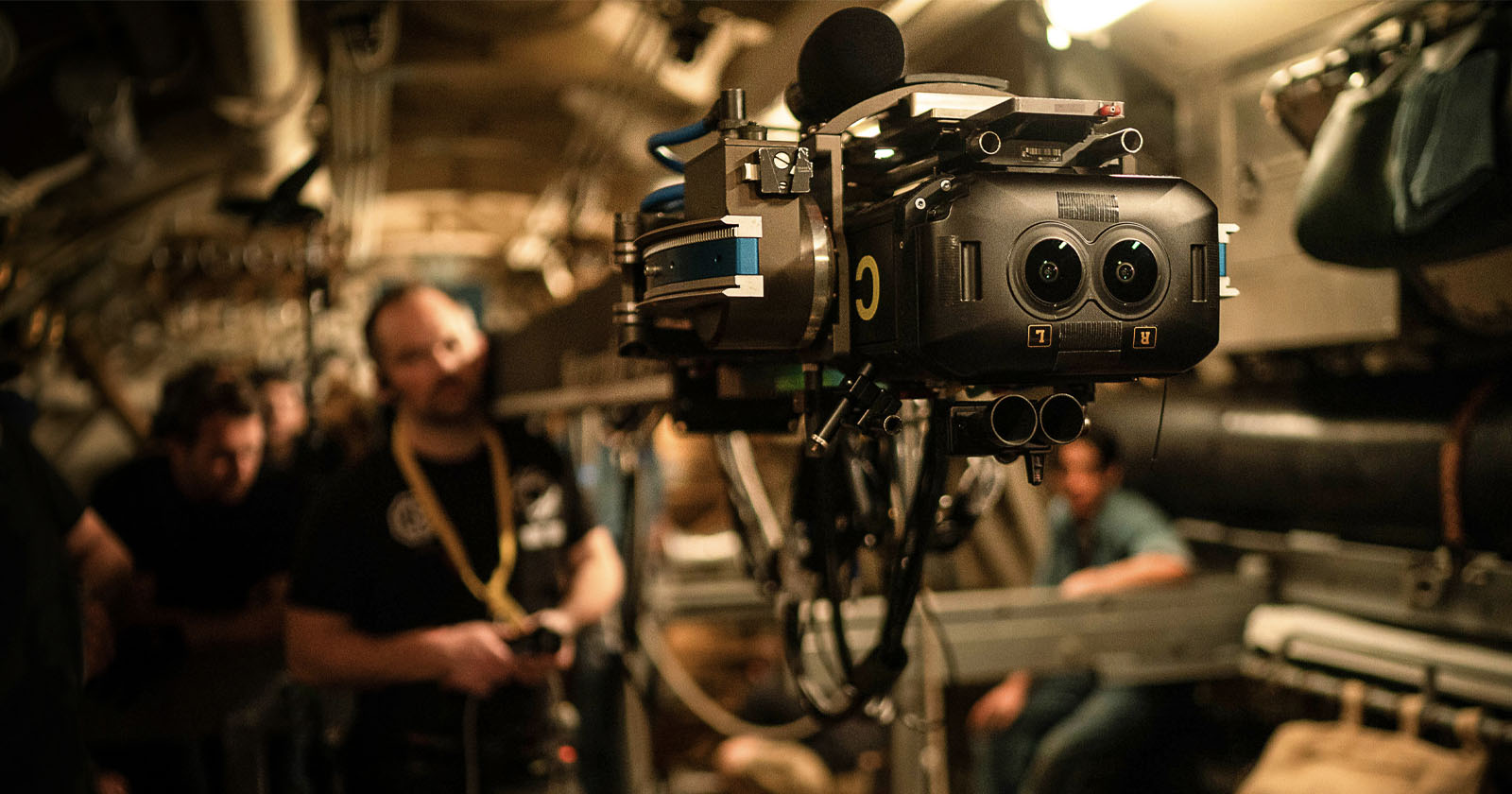 A professional camera rig is set up in a dimly lit industrial or mechanical environment. Several people are in the background, blurred and out of focus, suggesting the setting is a film shoot or a documentary setting. A professional camera rig is set up in a dimly lit industrial or mechanical environment. Several people are in the background, blurred and out of focus, suggesting the setting is a film shoot or a documentary setting.
