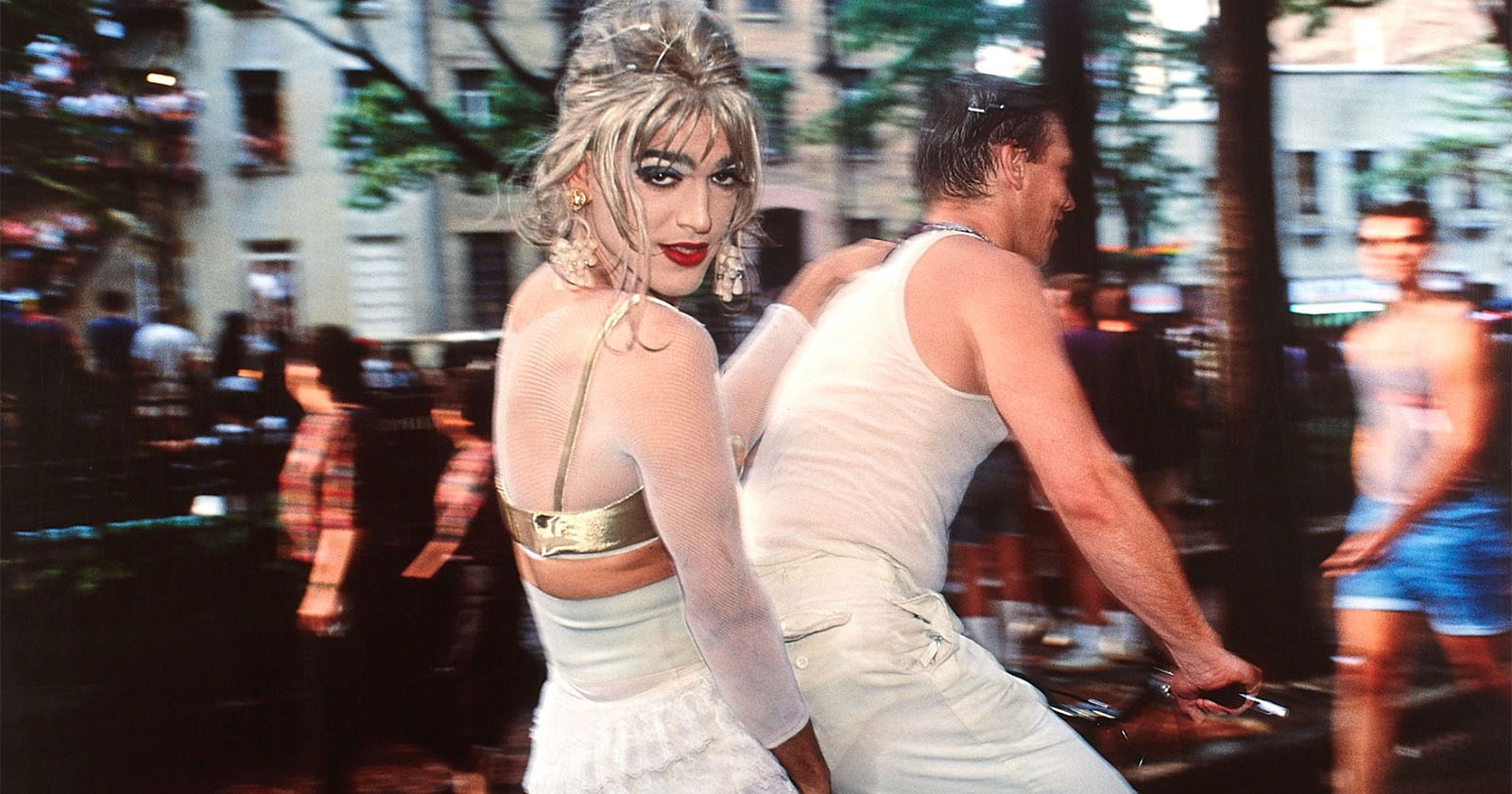 A person in a white outfit with blonde hair and red lipstick stands outdoors. They are alongside another person in a sleeveless white top riding a bicycle. The background is blurred with people and trees, suggesting a lively, urban scene. A person in a white outfit with blonde hair and red lipstick stands outdoors. They are alongside another person in a sleeveless white top riding a bicycle. The background is blurred with people and trees, suggesting a lively, urban scene.