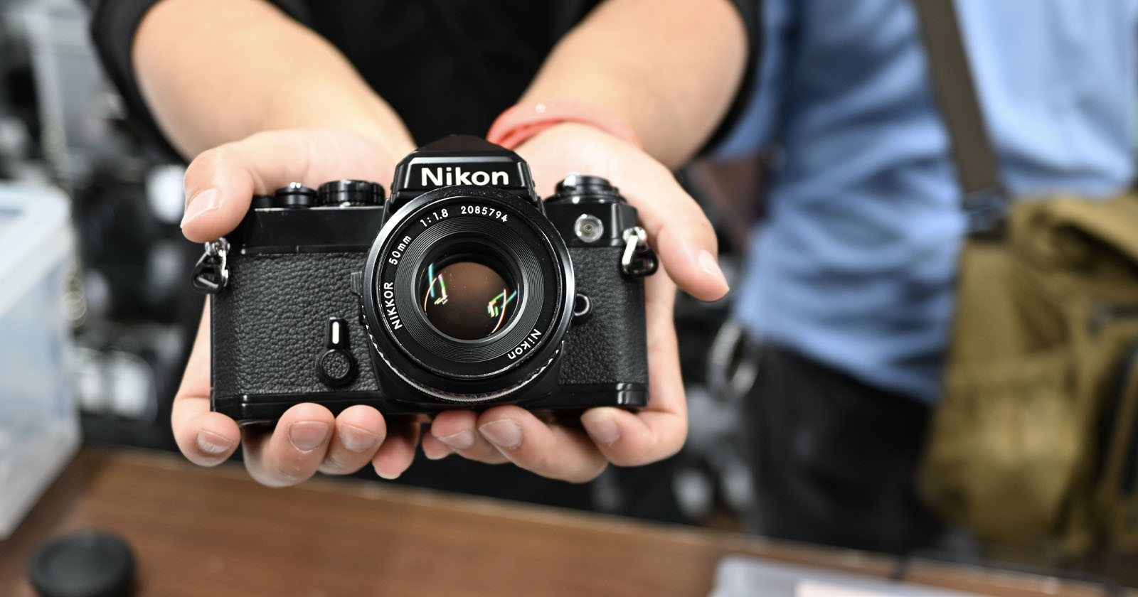 A person holds a vintage Nikon film camera towards the lens. The camera's body is black with visible details, including the Nikon logo and lens information. The background is slightly blurred, focusing attention on the camera. A person holds a vintage Nikon film camera towards the lens. The camera's body is black with visible details, including the Nikon logo and lens information. The background is slightly blurred, focusing attention on the camera.